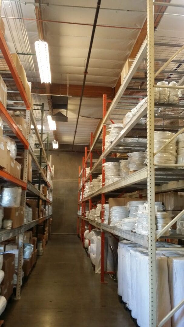 A warehouse aisle with tall metal shelves filled with stacked boxes and wrapped items. Fluorescent lights hang from the ceiling.