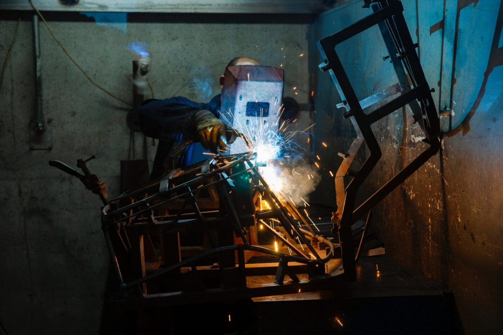 A person welds metal frames in a dimly lit workshop, with bright sparks flying from the welding process.