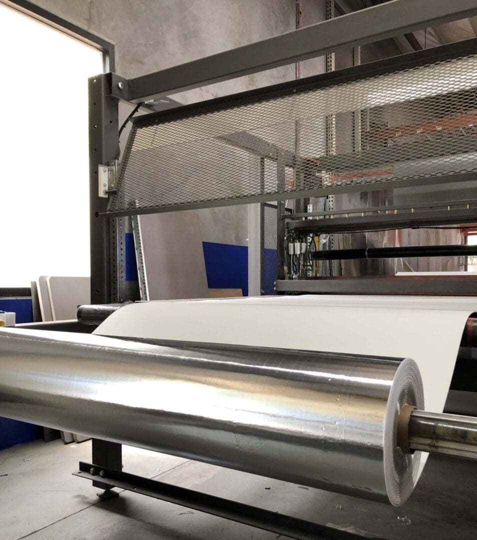 A large roll of white paper is unwinding on an industrial machine inside a factory setting.