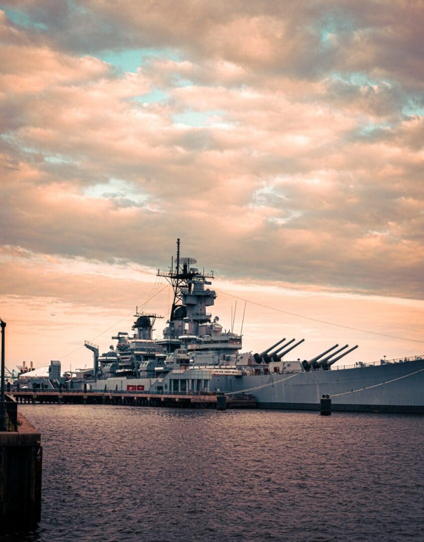 A large battleship docked at a harbor under a cloudy sky during sunset.