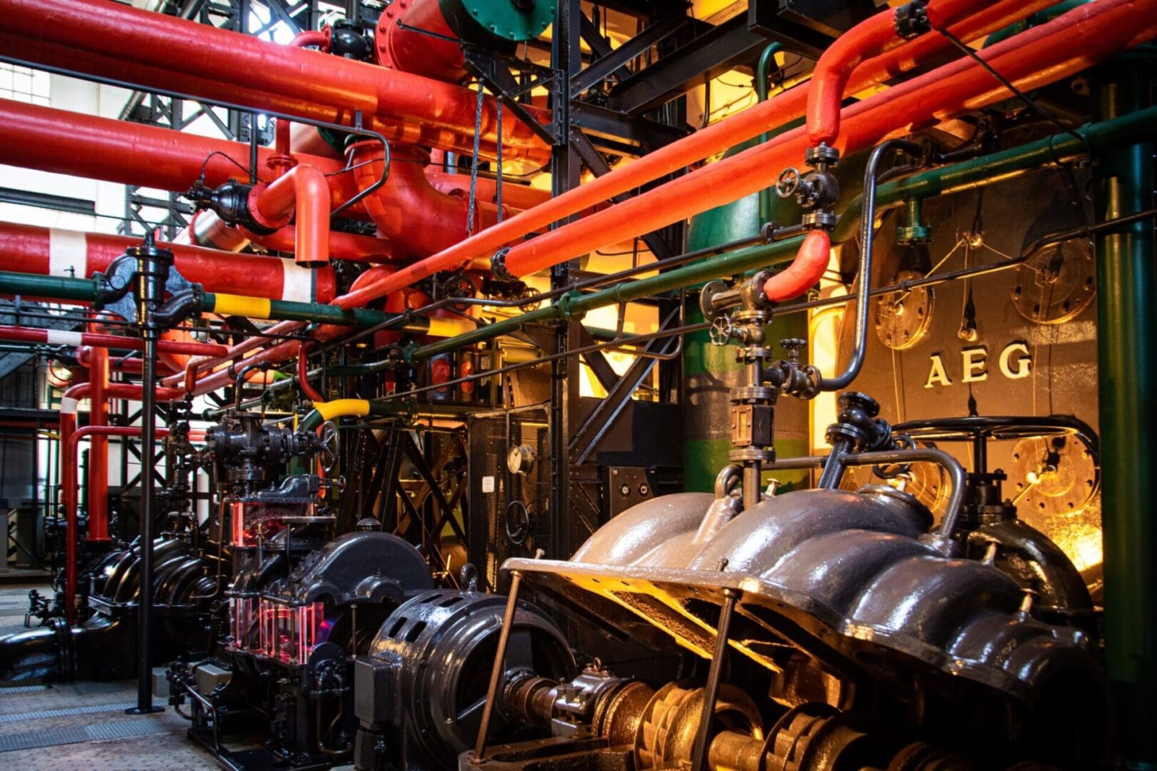 Industrial machinery with red and orange pipes and metallic structures in a factory setting. AEG logo visible on equipment.