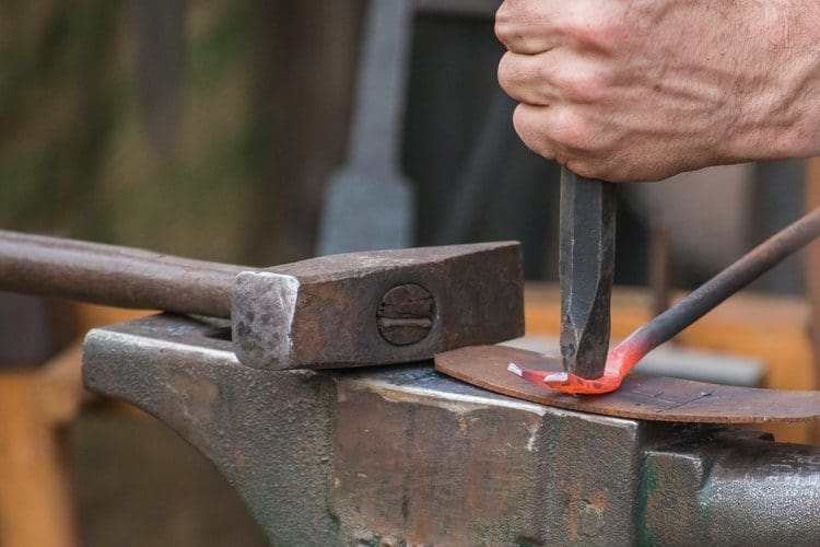Close-up of a person hammering a heated metal rod on an anvil, with a second tool for shaping the metal.