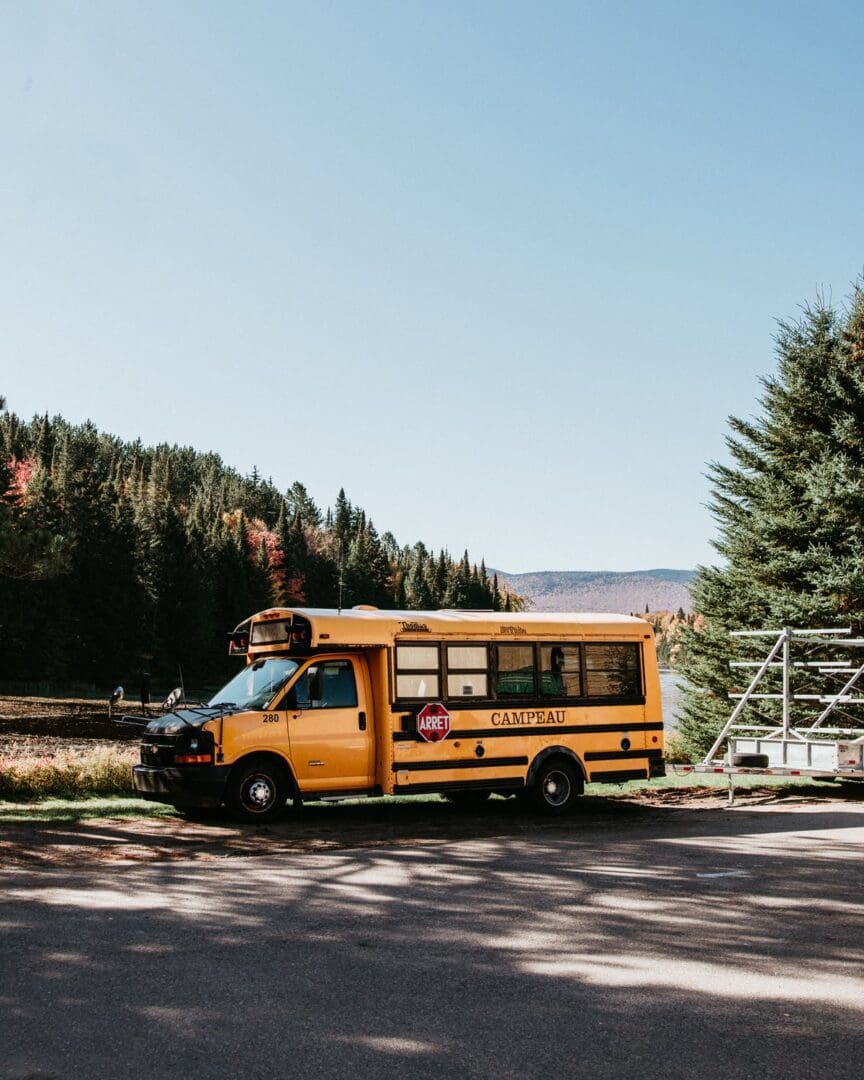 A small yellow school bus is parked on the side of a road near a lake, surrounded by trees and hills under a clear blue sky.