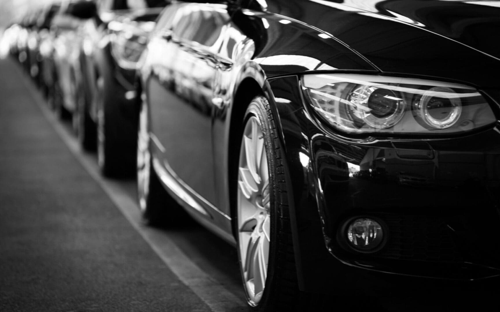 Close-up of the front end of a parked black car in a row of vehicles, highlighting the headlight and wheel, in a grayscale setting.