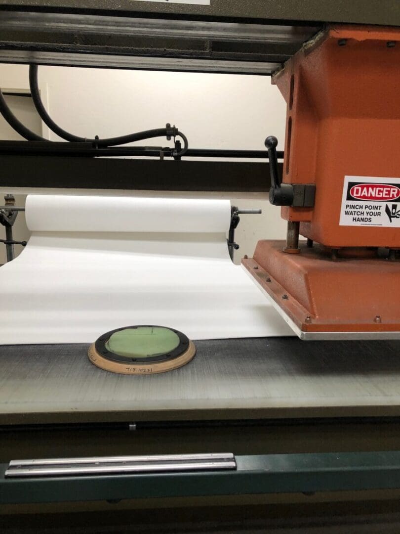 Industrial press machine with an orange casing and warning label, positioned over a roll of white material on a conveyor belt. A round object is placed on a plate beneath the press.