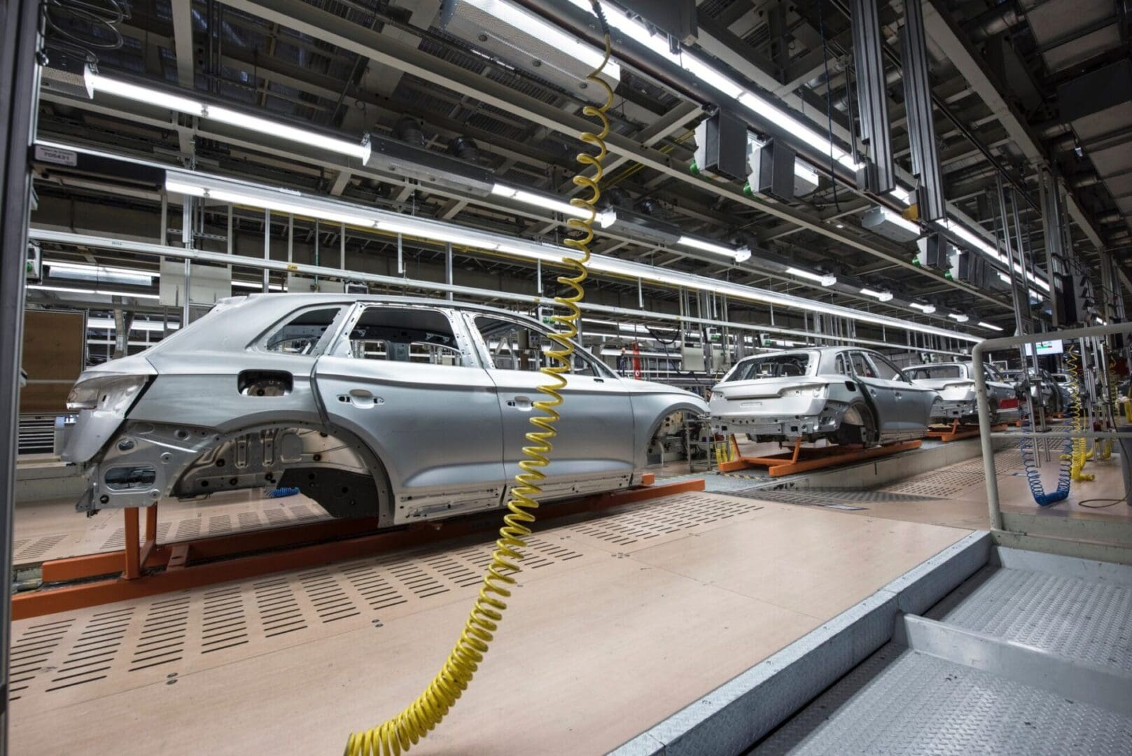 An automotive assembly line with unfinished car bodies on a conveyor. Bright overhead lights and suspended cables are visible.