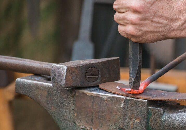 Close-up of a person hammering a heated metal rod on an anvil, with a second tool for shaping the metal.