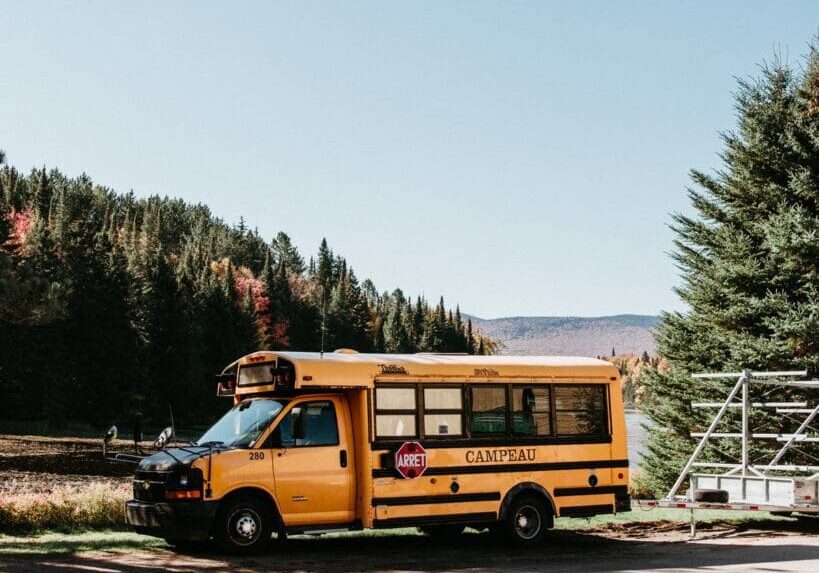 A small yellow school bus is parked on the side of a road near a lake, surrounded by trees and hills under a clear blue sky.