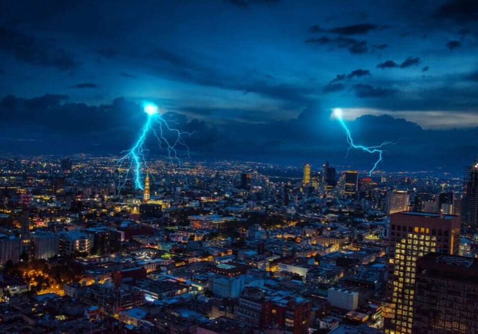A nighttime cityscape with two bright lightning strikes illuminating the sky, casting light over the dense urban area below.